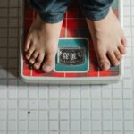 From above crop anonymous barefoot child in jeans standing on weigh scales on tiled floor of bathroom