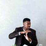 Businessman in a black blazer signaling time out with an hourglass and open book on a table.